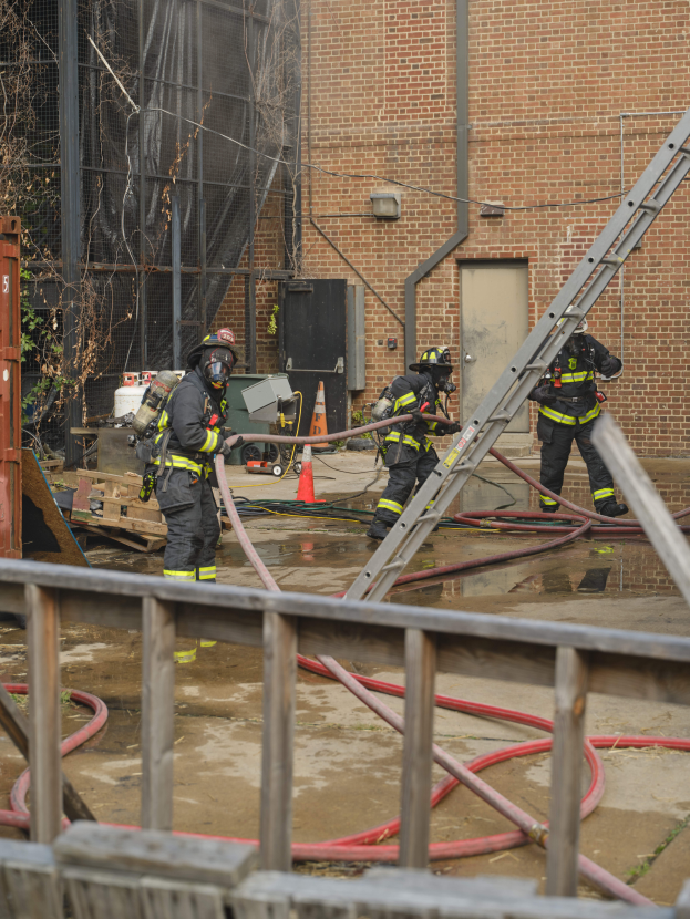 Feuerwehrleute in Helmen, die Rohre halten, arbeiten daran, ein Gebäude Feuer zu löschen, mit einem Metallzaun, verstreuten Rohren, einem Container, einem Verkehrskegel, verschiedenen Gegenständen, einem Gebäude mit Fenstern und einer Tür, einem Metallrahmen, Drähten, einem Baum und dem Himmel im Hintergrund.