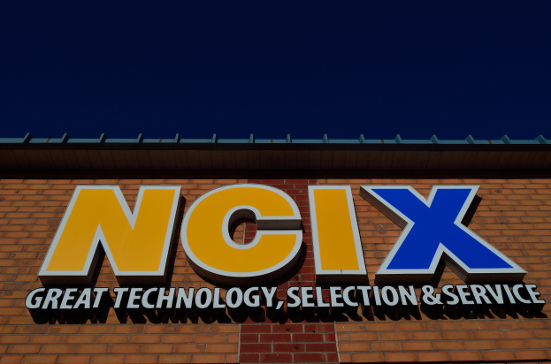 Ziegelgebäude mit einer Tafel, auf der "NCIX Great Technology, Selection & Service" steht, unter einem klaren blauen Himmel.