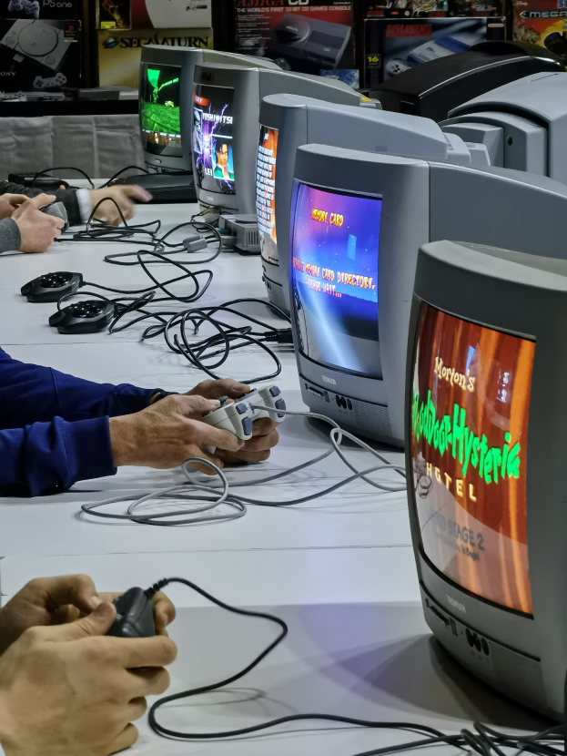 Eine Gruppe von Menschen sitzt um einen Tisch und spielt Videospiele, hält Controller, mit mehreren Fernsehern und Kabeln auf dem Tisch und Postern an der Wand im Hintergrund.