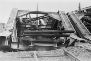 Ein Schwarz-Weiß-Foto einer Schienenstraße, die zu einer baufälligen Brücke mit kaputten Geländern und Schutt führt, umgeben von Gebäuden unter einem klaren Himmel.