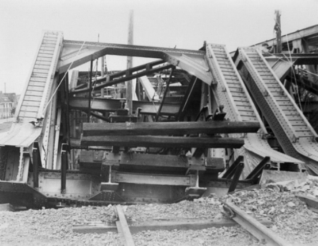Ein Schwarz-Weiß-Foto einer Schienenstraße, die zu einer baufälligen Brücke mit kaputten Geländern und Schutt führt, umgeben von Gebäuden unter einem klaren Himmel.