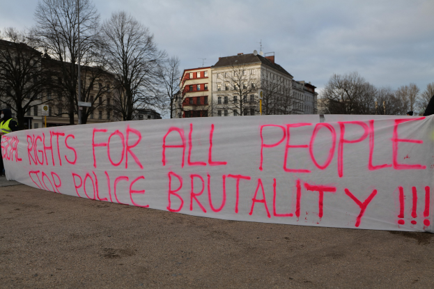 Gruppe von Menschen mit einem Banner mit der Aufschrift "Rechte für alle Menschen Stoppt Polizeigewalt" vor einem Laternenpfahl, einem Schild, Bäumen, Gebäuden mit Fenstern und einem bewölkten Himmel.