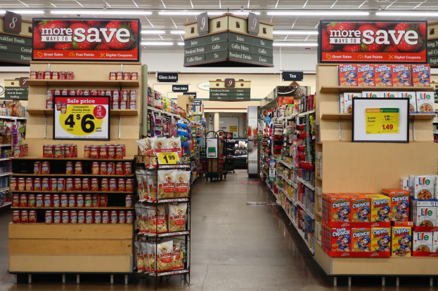 Ein Innenraum eines Lebensmittelladens mit Regalen und Bodenauslagen, die mit verschiedenen Lebensmitteln bestückt sind, Deckenbeleuchtung und einer nummerierten Tafel.