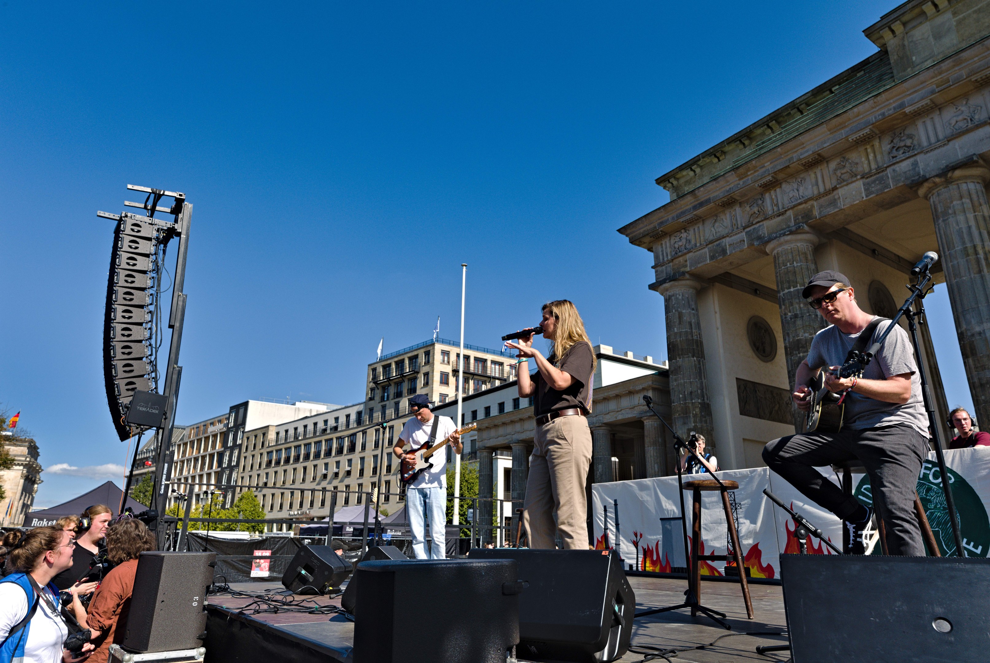 Eine Gruppe von Menschen, die auf einer Bühne vor dem Brandenburger Tor in Berlin Musik spielen, mit Lautsprechern und Equipment im Freien, vor einem klaren blauen Himmel.