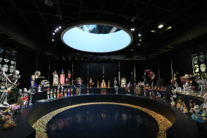 Ein großer Raum voller verschiedener Spielzeuge, darunter Skelette, die auf dem Boden angeordnet sind, mit Schädeln an den Wänden und Lampen an der Decke, die eine festliche Atmosphäre für den Tag der Toten schaffen.