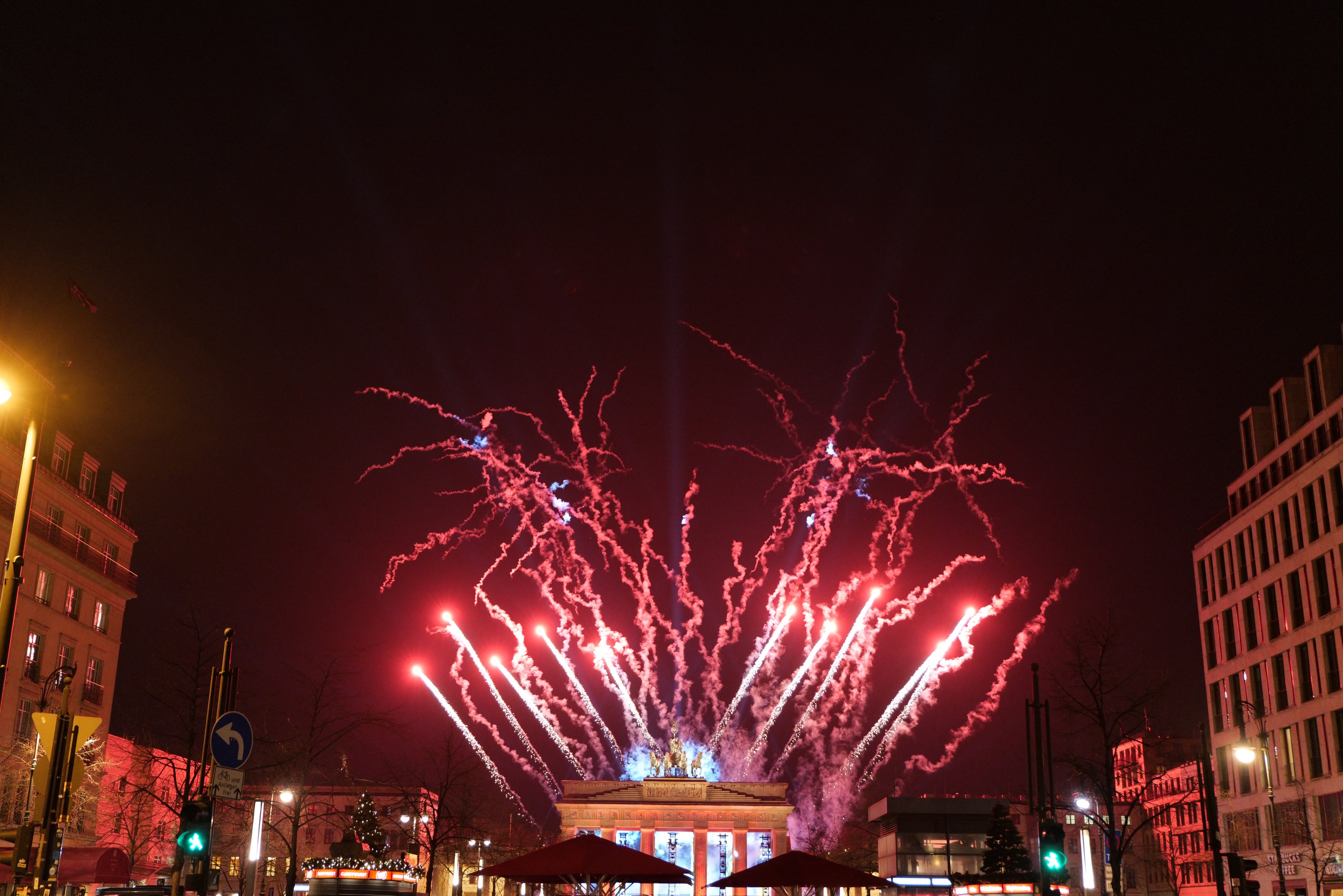 Eine belebte Straße in Berlin öffnet sich am Silvesterabend, mit Menschen, Fahrzeugen, Gebäuden und Feuerwerk, das den Nachthimmel erhellt.