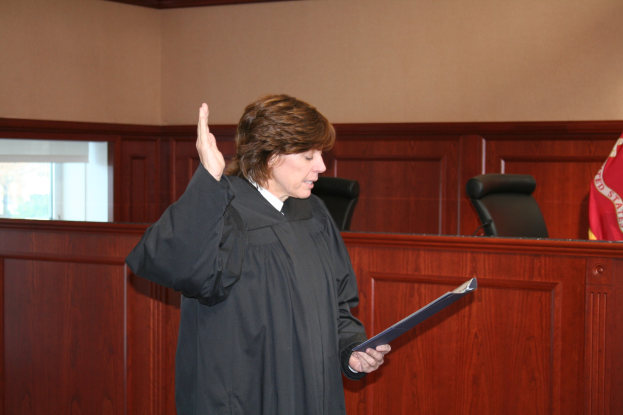 Eine Frau in einer schwarzen Robe steht vor einem Richtertisch, hält eine Akte und ein Mikrofon und eine Flagge auf dem Tisch und Stühle und ein Fenster dahinter.