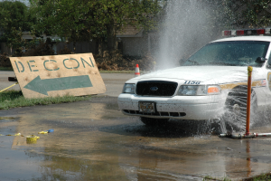 Ein Polizeiauto fährt durch eine Wasserlache, mit einer Tafel, einem Verkehrskegel und anderen Gegenständen am Boden, mit Bäumen, Gebäuden und einem klaren blauen Himmel im Hintergrund.