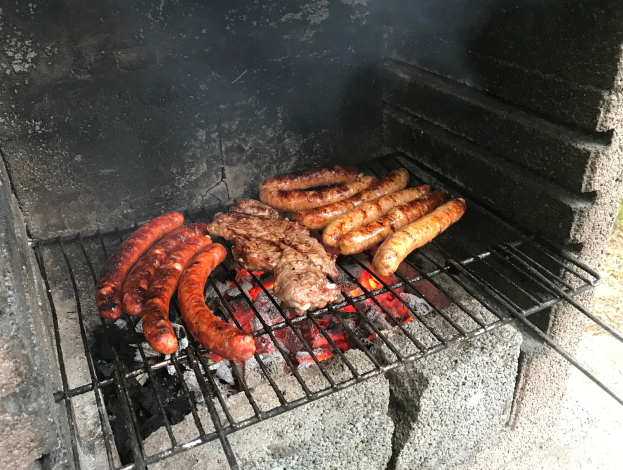 Ein Grill mit Würstchen und Fleisch darauf, umgeben von Kohle und Feuer, auf Gras im Freien