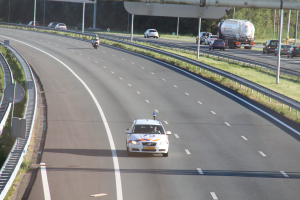 Ein Auto fährt auf einer Autobahn mit einem Polizeiwagen am Straßenrand, anderen Fahrzeugen, Geländern, Pfosten, Planken und Bäumen im Hintergrund.