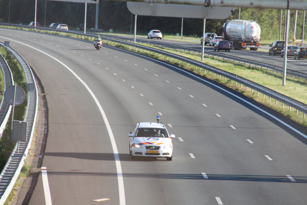 Ein Auto fährt auf einer Autobahn mit einem Polizeiwagen am Straßenrand, anderen Fahrzeugen, Geländern, Pfosten, Planken und Bäumen im Hintergrund.