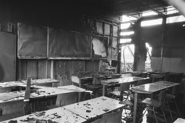 Schwarz-weißes Foto eines Klassenzimmers mit Tischen, Stühlen, verkohlten Wänden und zerbrochenen Fenstern, das eine trostlose und verlassene Atmosphäre vermittelt.