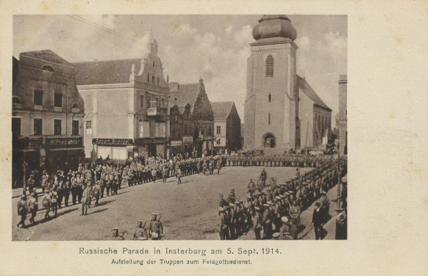 Ein Schwarz-Weiß-Foto eines Umzugs in Insterburg am 5. September 1914 mit vielen Menschen, Gebäuden und bewölktem Himmel sowie Text unten.