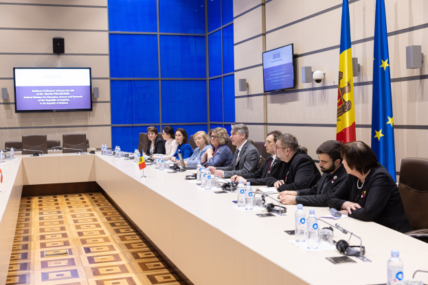 Eine Gruppe von Menschen, die um einen langen Tisch sitzen, mit Flaschen, Gl├Ąsern, Mikrofonen, Papieren, Flaggen, Fernsehern und Lautsprechern.