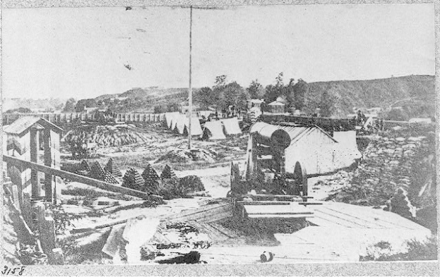 Schwarz-weißes Foto einer Baustelle mit einer Maschine im Vordergrund, Schuppen, Bäumen, Hügel und Himmel im Hintergrund, mit Text unten, der "Die Bauarbeiten für eine neue Brücke in der Stadt San Francisco, Kalifornien" lautet.