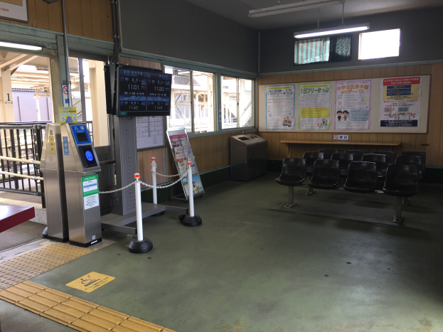 Ein belebter Bahnhofseingang mit Sitzgelegenheiten, einem Service-Schalter, Ticketautomaten, Kettenabsperrungen, Informationsanzeigen, Wandplakaten, Deckenleuchten und Geländern.