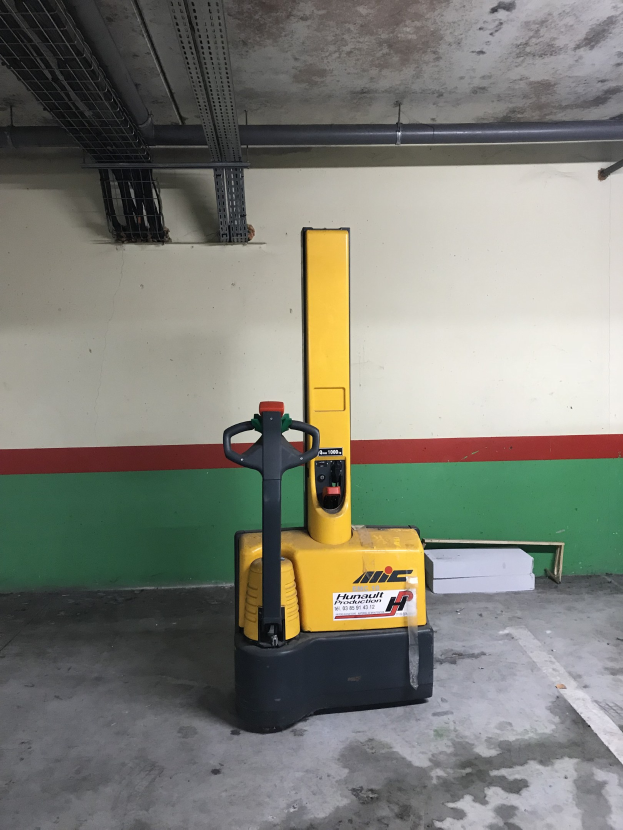 Ein gelber Gabelstapler steht in einem Lager mit Metallregalen auf der linken Seite und einer Wand im Hintergrund.