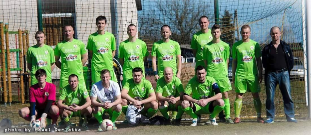 Eine Gruppe von Männern in grünen T-Shirts steht auf einem Fußballfeld, einige sind in der Hocke, mit einem Ball vor ihnen und einem Torpfosten rechts daneben, Bäume und Gebäude im Hintergrund.