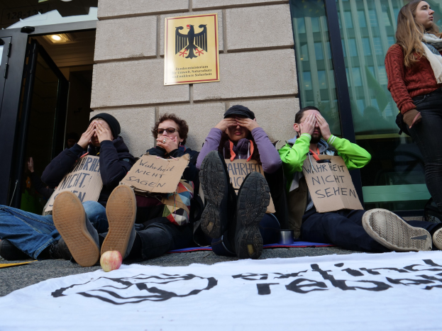 Eine Gruppe von Menschen, die vor einem Gebäude auf dem Boden sitzen, Schilder halten und ein Banner auf der Straße sowie eine Frau rechts daneben zu sehen; das Gebäude hat Glasfenster und eine Tür, und in Hintergrund ist eine Wand mit einer Tafel zu erkennen.