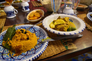 Ein Tisch mit Tellern, Gläsern, Schüsseln, Krügen, Bestecken und Matten, mit im Hintergrund sitzenden Menschen bei einem traditionellen marokkanischen Essen.