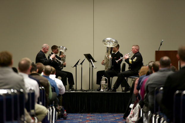 Eine Gruppe von Männern, die auf einer Bühne Brass-Instrumente spielen, mit Notenständen, einem Podium mit Mikrofon und einem Publikum, das auf Stühlen sitzt.