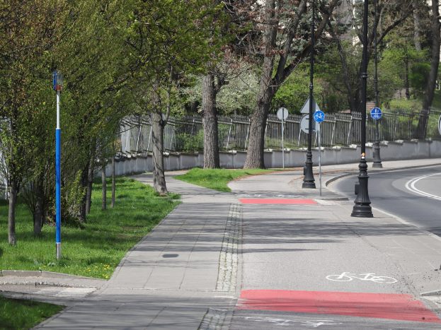 Stadtstraße mit einem ausgewiesenen Fahrradweg, gesäumt von Bäumen, Schildern und einem Zaun, umgeben von Gras und Pflanzen, mit Gebäuden und einem klaren blauen Himmel im Hintergrund.