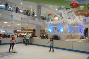 Eine Gruppe von Menschen, die auf einer Eisbahn in einem Einkaufszentrum Schlittschuh laufen, umgeben von einem häuslichen Aufbau, der mit Lichtern und festlichen Dekorationen geschmückt ist, mit Zuschauern und zusätzlichen Dekorationen, die eine festliche Atmosphäre schaffen.