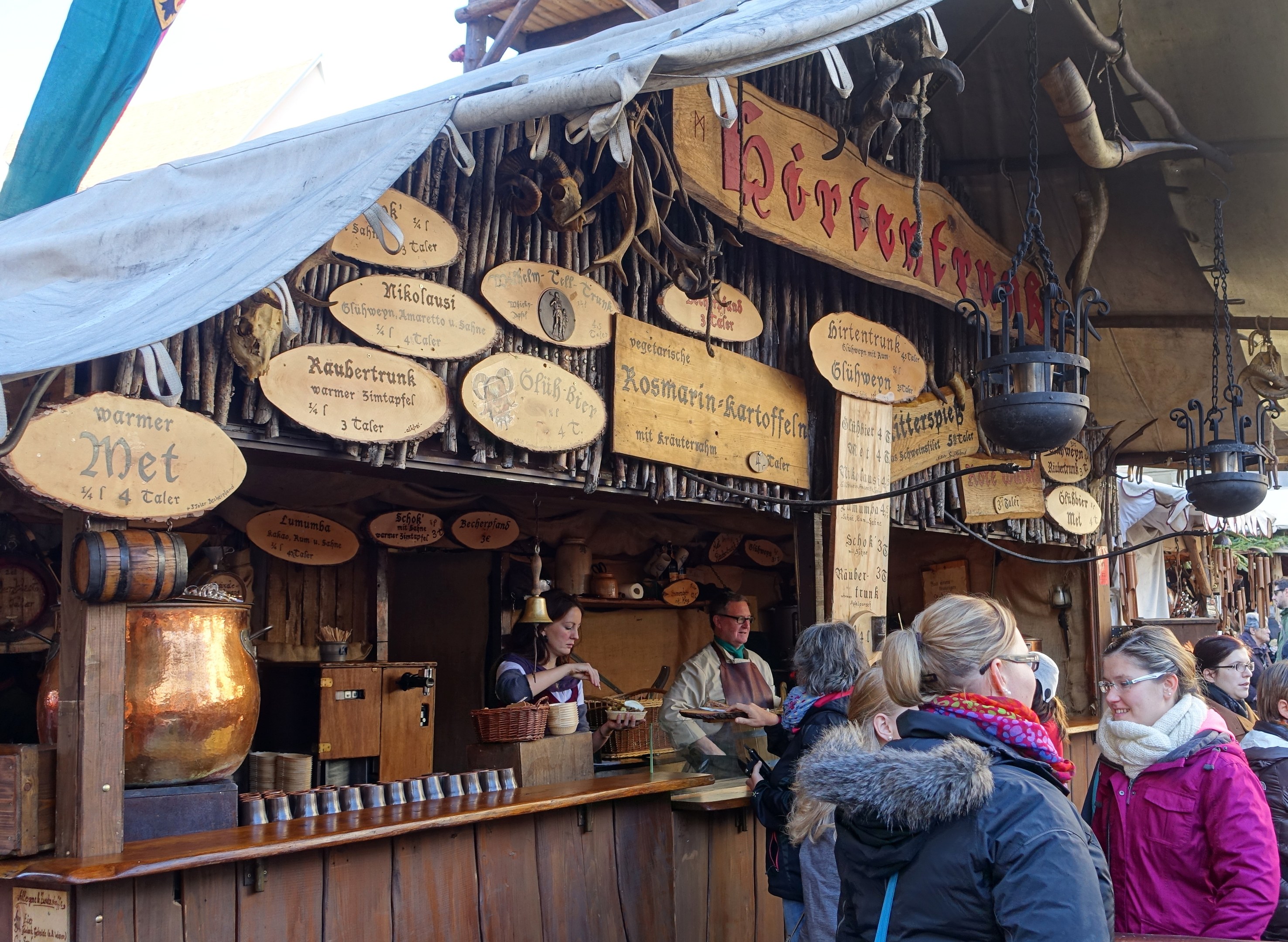 Eine Gruppe von Menschen steht vor einem Holzstand auf einem Outdoor-Festival, mit Brettern, Töpfen und Laternen auf dem Stand und Bäumen und einem klaren Himmel im Hintergrund.