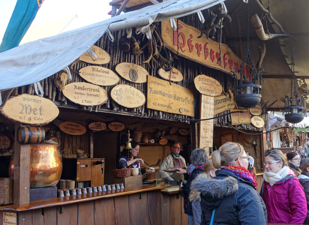 Eine Gruppe von Menschen steht vor einem Holzstand auf einem Outdoor-Festival, mit Brettern, Töpfen und Laternen auf dem Stand und Bäumen und einem klaren Himmel im Hintergrund.