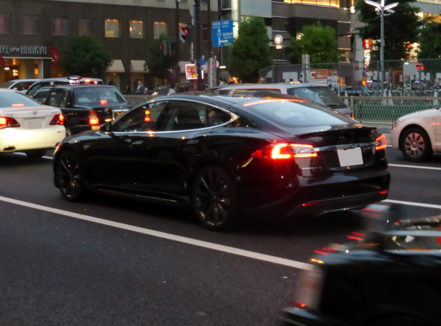 Schwarzer Tesla Model S fährt nachts durch eine Stadtstraße mit anderen Fahrzeugen, Gebäuden, Bäumen, Laternen, Verkehrssignalen, Schildern, Geländern und Passanten im Hintergrund.