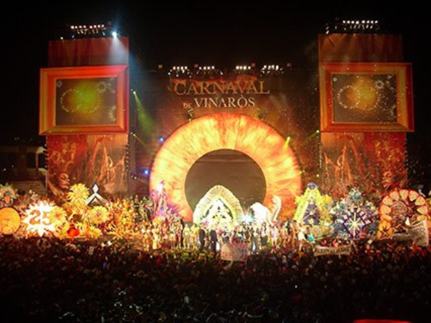 Eine Menschenmenge bei einem nächtlichen Karneval mit beleuchteten Fahrgeschäften, Pfählen und Bäumen unter einem bewölkten Himmel.