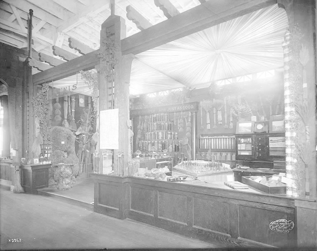 Altes Schwarz-Weiß-Foto eines Apothekeninneren mit einem zentralen Tresen, der verschiedene Gegenstände zeigt, Regalen im Hintergrund mit Flaschen und Artikeln und Text unten, der darauf hinweist, dass es sich um eine Apotheke handelt.