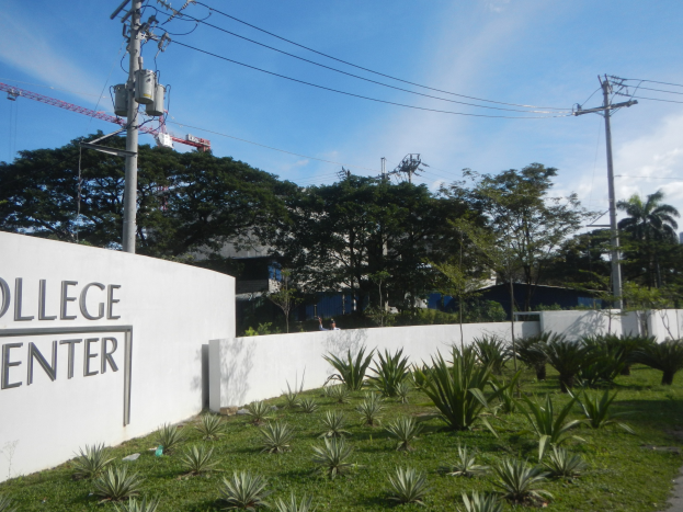 Eingang zu einem College-Zentrum mit einem "College-Zentrum"-Schild, umgeben von Grünflächen, einem Weg, Strommasten, Bäumen, Gebäuden, einem Kran und einem bewölkten Himmel.