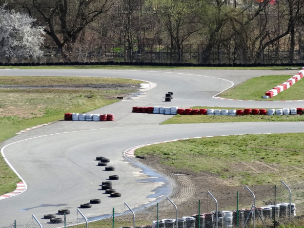 Rennstrecke mit mehreren gestapelten Reifen an den Seiten, umgeben von Gras und Maschendrahtzaun, mit Bäumen im Hintergrund.