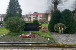 Ein Denkmal in einem Park mit einem Kranz, umgeben von Gras, Pflanzen, Blumen und Bäumen, gewidmet den Opfern des Zweiten Weltkriegs, mit Gebäuden und Himmel im Hintergrund.