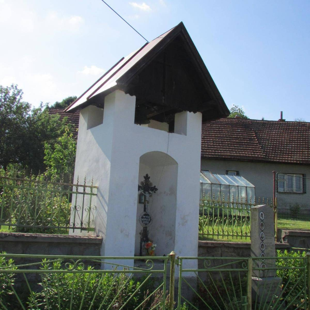 Kleines weißes Kirchengebäude mit einem Kreuz darauf, umgeben von einem Metallzaun, Pflanzen, Gras, einem Weg, Häusern, Polen, Drähten und einem bewölkten Himmel, identifiziert als die Kirche des Heiligen Grabes.