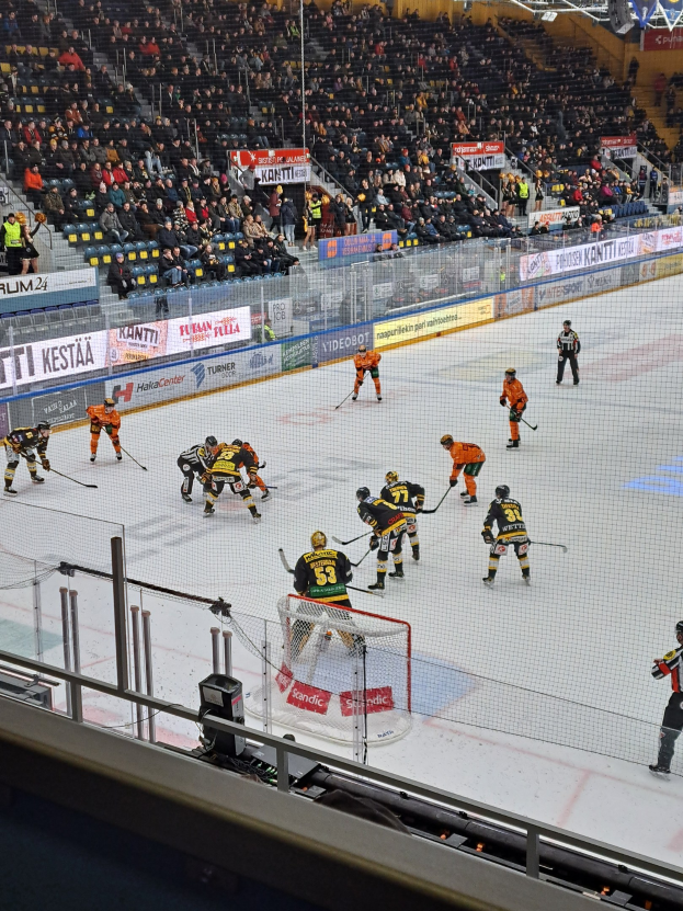 Gruppe von Menschen, die Hockey auf einer Indoor-Eisfläche mit Netz und Umzäunung spielen, mit Zuschauern im Hintergrund, Bannern und Überdachungsbeleuchtung