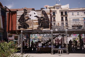 Menschen sitzen und stehen vor einem Graffiti-bedeckten Gebäude mit Bänken, Pflanzen und urbanen Elementen im Hintergrund.
