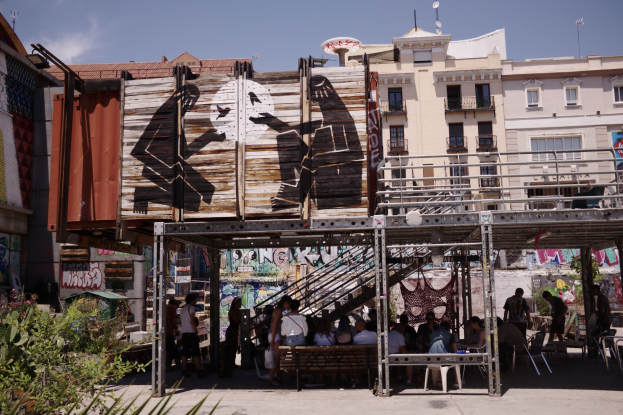 Menschen sitzen und stehen vor einem Graffiti-bedeckten Gebäude mit Bänken, Pflanzen und urbanen Elementen im Hintergrund.