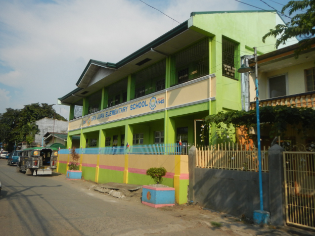 Ein Schulgebäude mit einer grünen und gelben Fassade, umgeben von einer Straße mit Fahrzeugen, Topfpflanzen, einem Zaun, einem Tor, einem Laternenmast, Bäumen, Drähten und einem bewölkten Himmel.