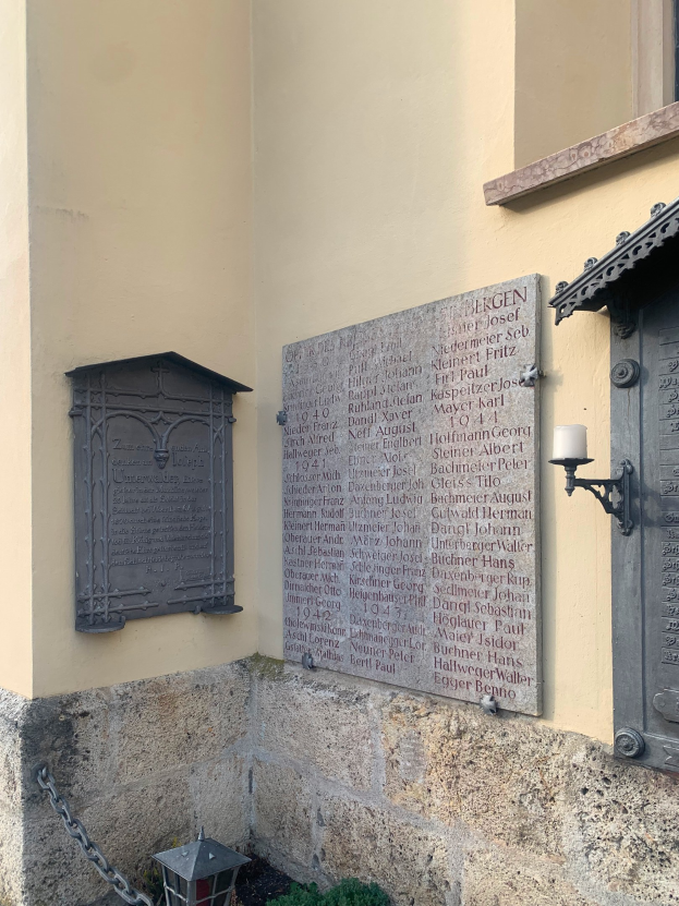 Ein Gebäude mit einer Holocaust-Gedenktafel, einem Fenster, einer Tür, einer Äußenbeleuchtung, einer Kette und umgebenden Pflanzen.