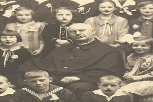 Ein altes Schwarz-Wei├č-Foto eines strengen Mannes, der in der Mitte sitzt und von einer Gruppe von Kindern umgeben ist, die alle bewundernd zu ihm aufschauen.