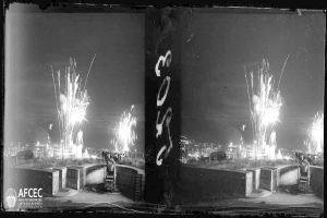 Ein Schwarz-Weiß-Foto von Feuerwerk, das über einer Stadt explodiert, bei der Catalunya-Feuerwerksfest in Mexiko-Stadt, Mexiko im Jahr 1939, mit Menschen und Gebäuden unten.