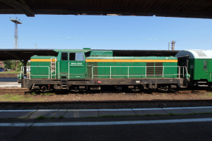 Ein grüner Diesel-Elektrolok auf Eisenbahnschienen unter einer Brücke, umgeben von Steinen, Gras, einem Weg, Bäumen, Strommasten mit Drähten, einem Turm, Fahrzeugen und einem bewölkten Himmel.