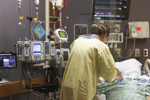 Ein Mann liegt in einem Krankenhausbett neben einem Monitor, mit einer Frau neben ihm; ein Tisch mit einem Monitor ist links, und verschiedene medizinische Geräte sind im Hintergrund.