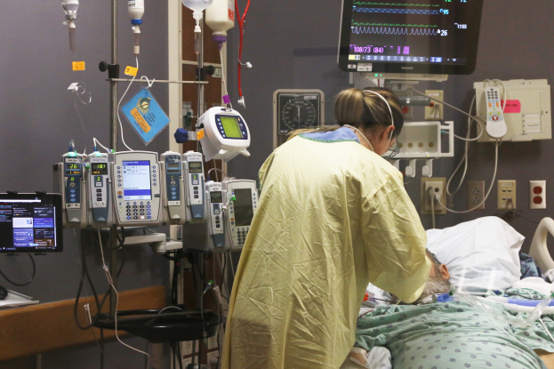 Ein Mann liegt in einem Krankenhausbett neben einem Monitor, mit einer Frau neben ihm; ein Tisch mit einem Monitor ist links, und verschiedene medizinische Geräte sind im Hintergrund.