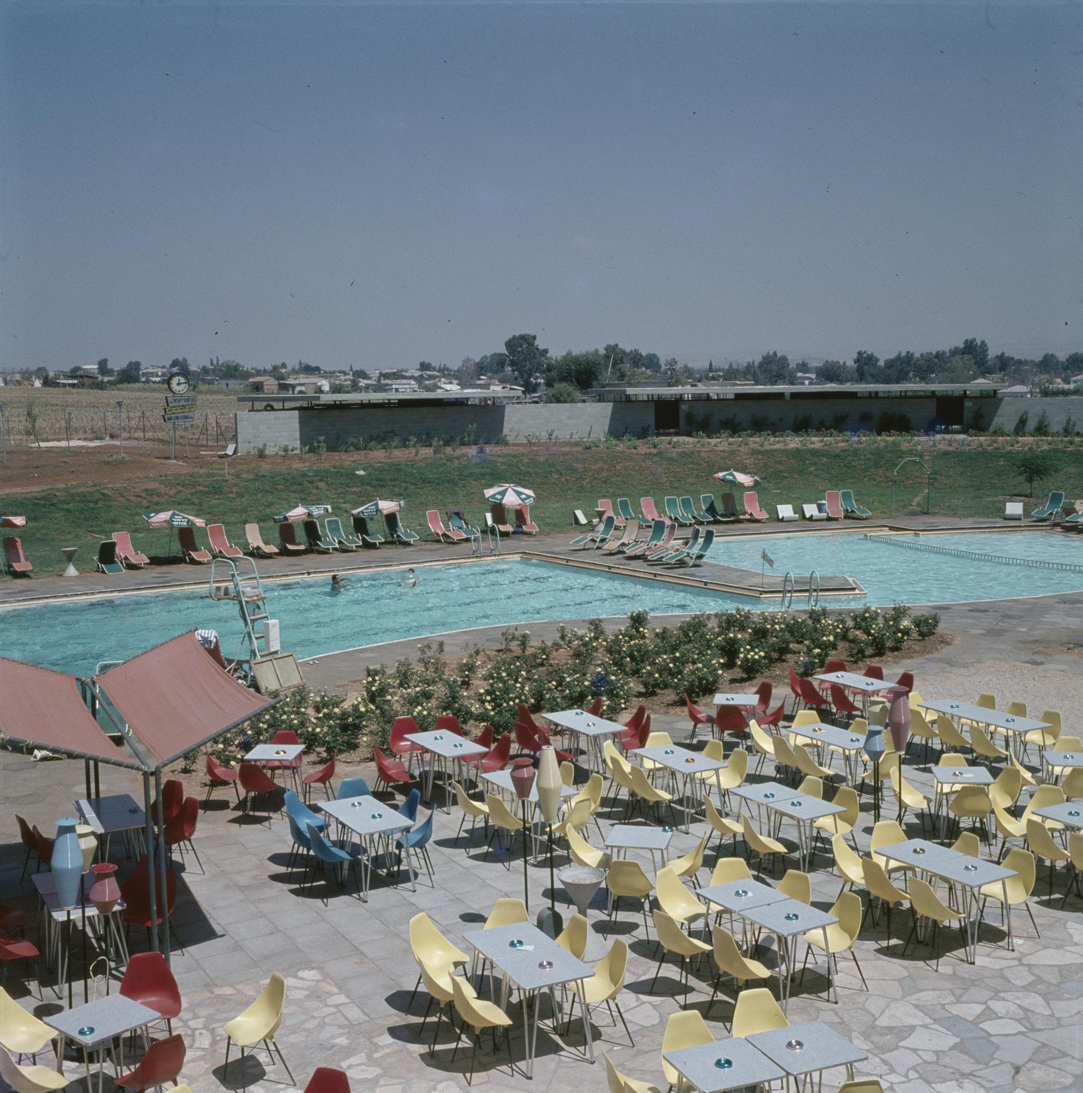 Ein Freibad umgeben von Tischen, Stühlen, Blumenpflanzen, Sonnenschirmen, Pfählen, einem Zaun, einer Wand, einer Baumgruppe, einigen Gebäuden, unter einem bewölkten Himmel.