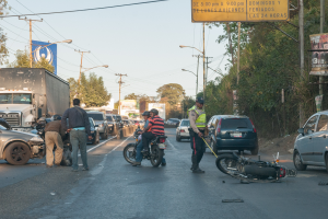 Eine Gruppe von Menschen steht um ein verunglücktes Motorrad auf der Straße mit mehreren Fahrzeugen, darunter ein Lastwagen, und einer Hintergrundlandschaft aus Bäumen, Pfählen, Laternen und Schildern unter dem Himmel.