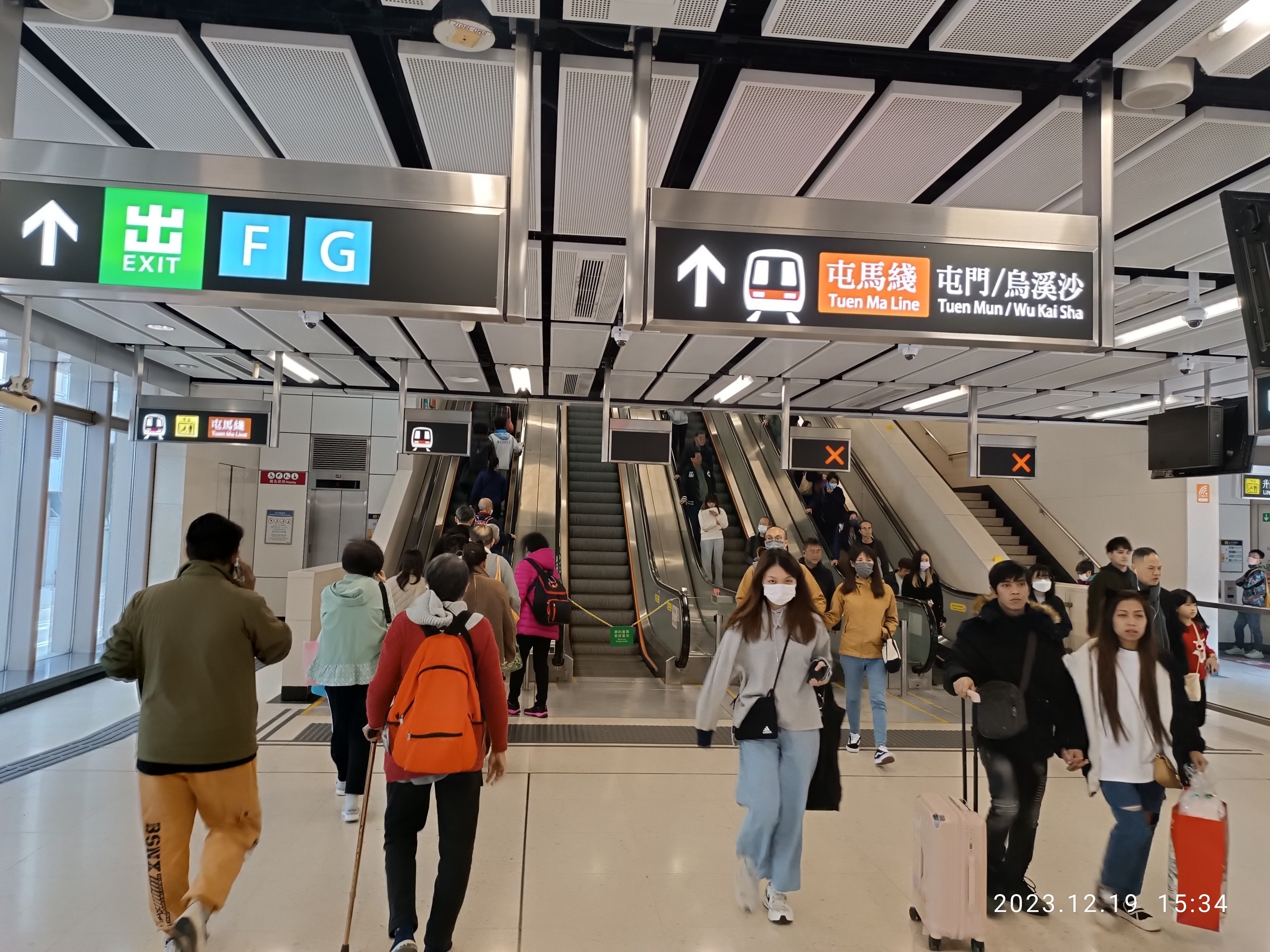 Menschen mit Gepäck durch einen geschlossenen Flughafen, Rolltreppen und Informationsanzeigen im Hintergrund.