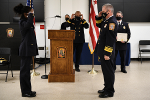 Gruppe uniformierter Personen in Masken vor einem Rednerpult mit Mikrofon bei einer Amtseinführungszeremonie für einen neuen Feuerchef, umgeben von Fahnen, Stühlen, Tischen und einer Wand.